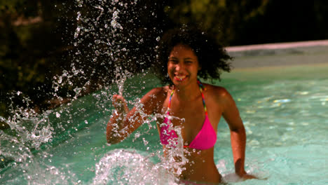 happy woman in pink bikini splashing at camera in swimming pool