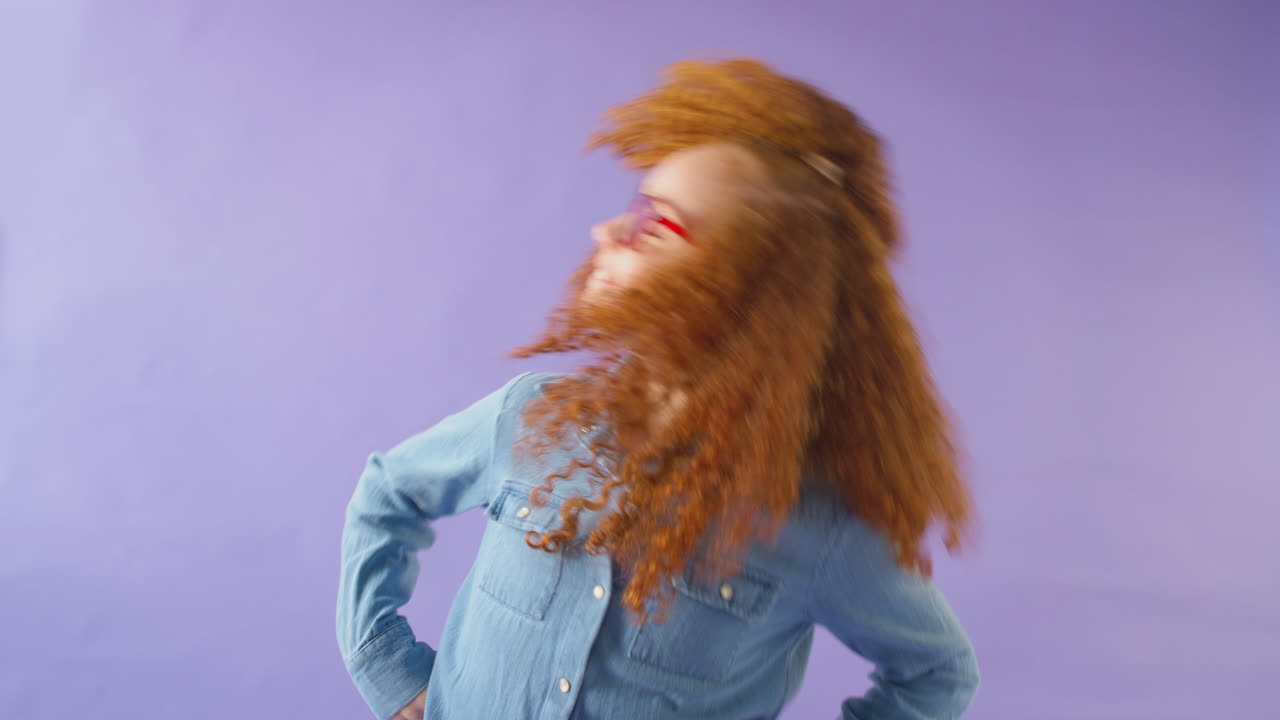 빨간 머리와 안경을 입은 어린 소녀의 스튜디오 이 보라색 배경에 머리를 흔들고 있습니다.