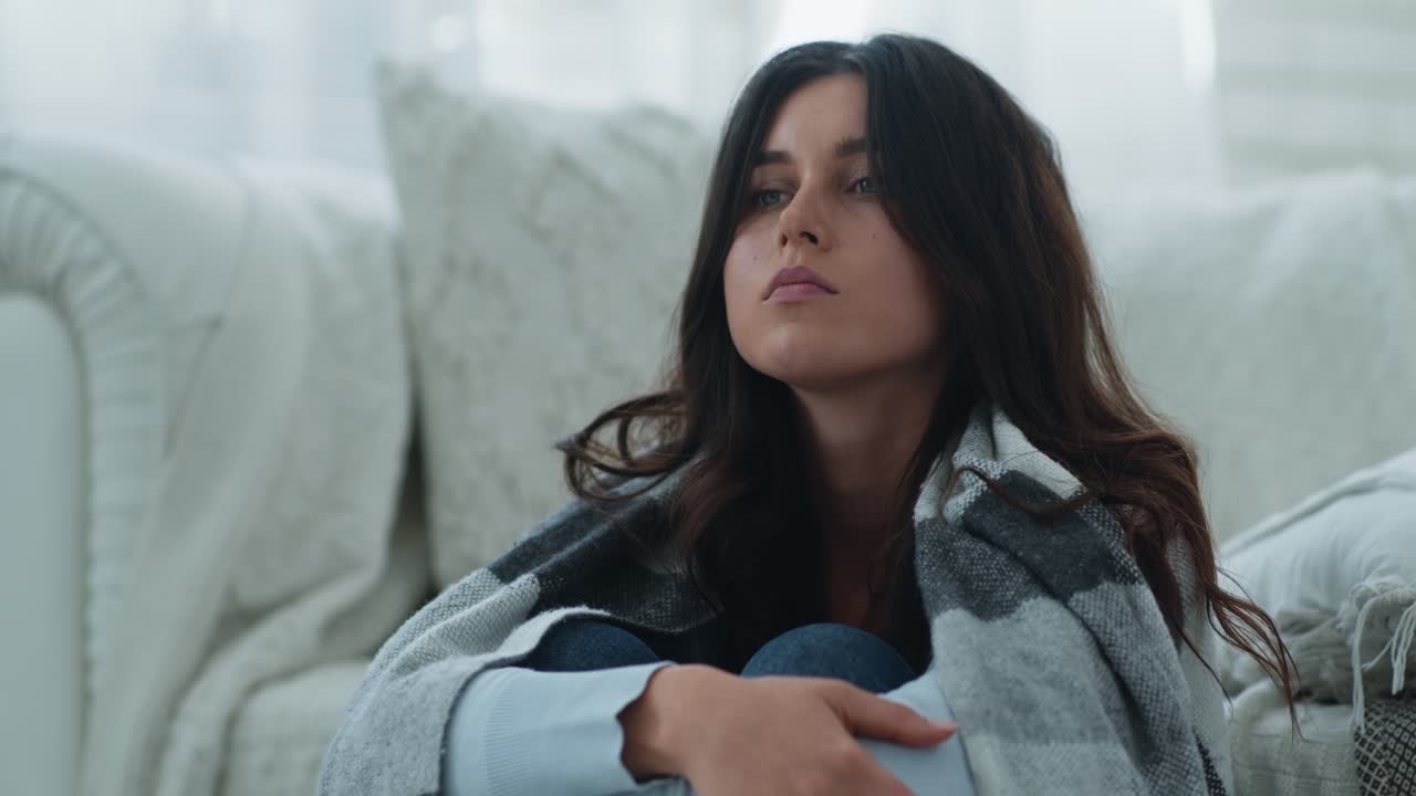 A young woman sitting on a sofa, wrapped in a blanket, looking thoughtful and sad.