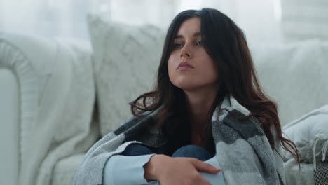a young woman sitting on a sofa, wrapped in a blanket, looking thoughtful and sad.