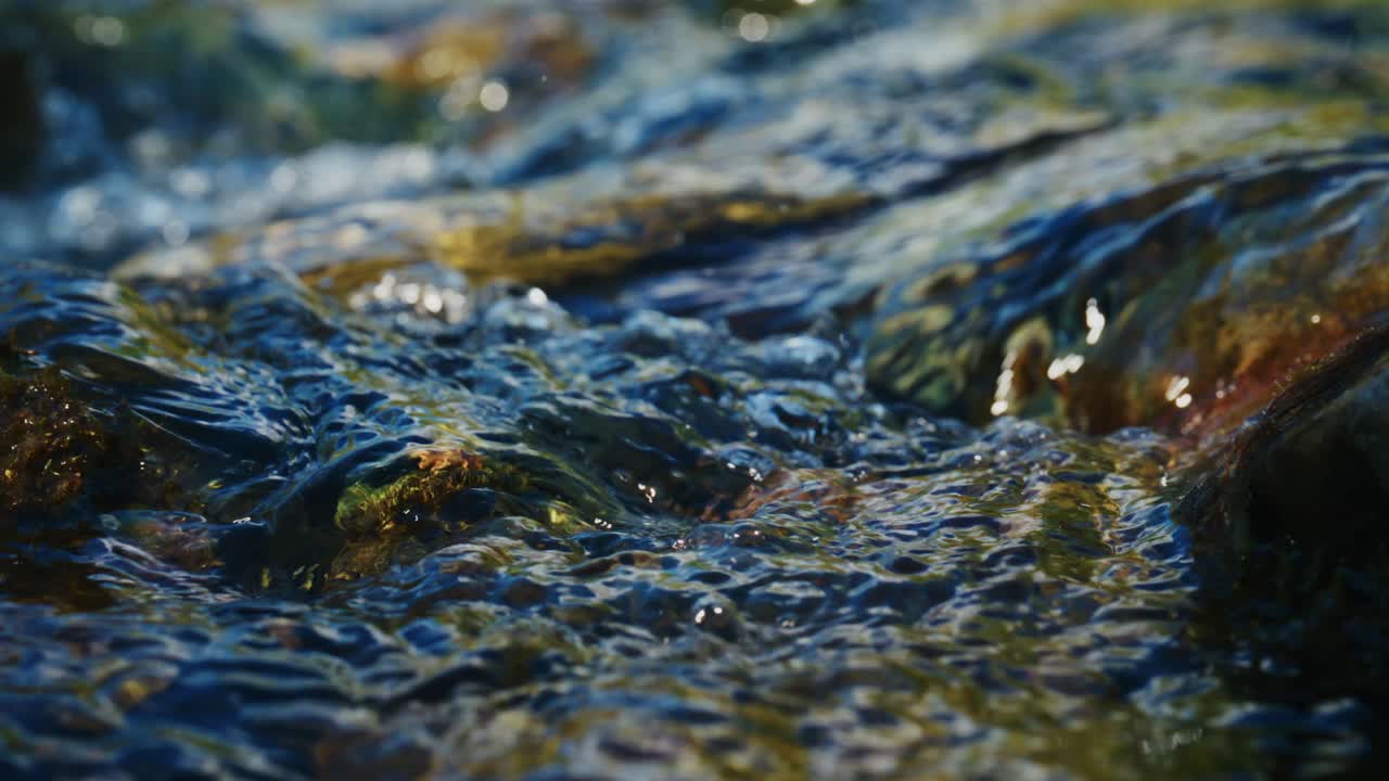 Primo piano di acqua limpida e impetuosa che scorre su rocce lisce e muschiose. La luce del sole scintilla su increspature e goccioline, creando riflessi scintillanti. Il movimento dell'acqua suggerisce energia naturale e freschezza.
