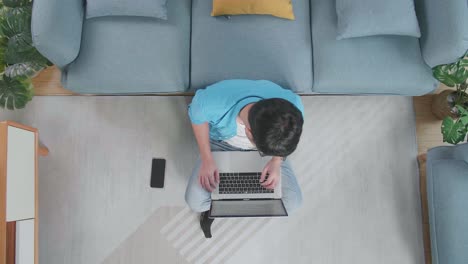top view zoom out shot of a tired man having a headache while typing on a laptop on the floor with smartphone in front of a sofa at home