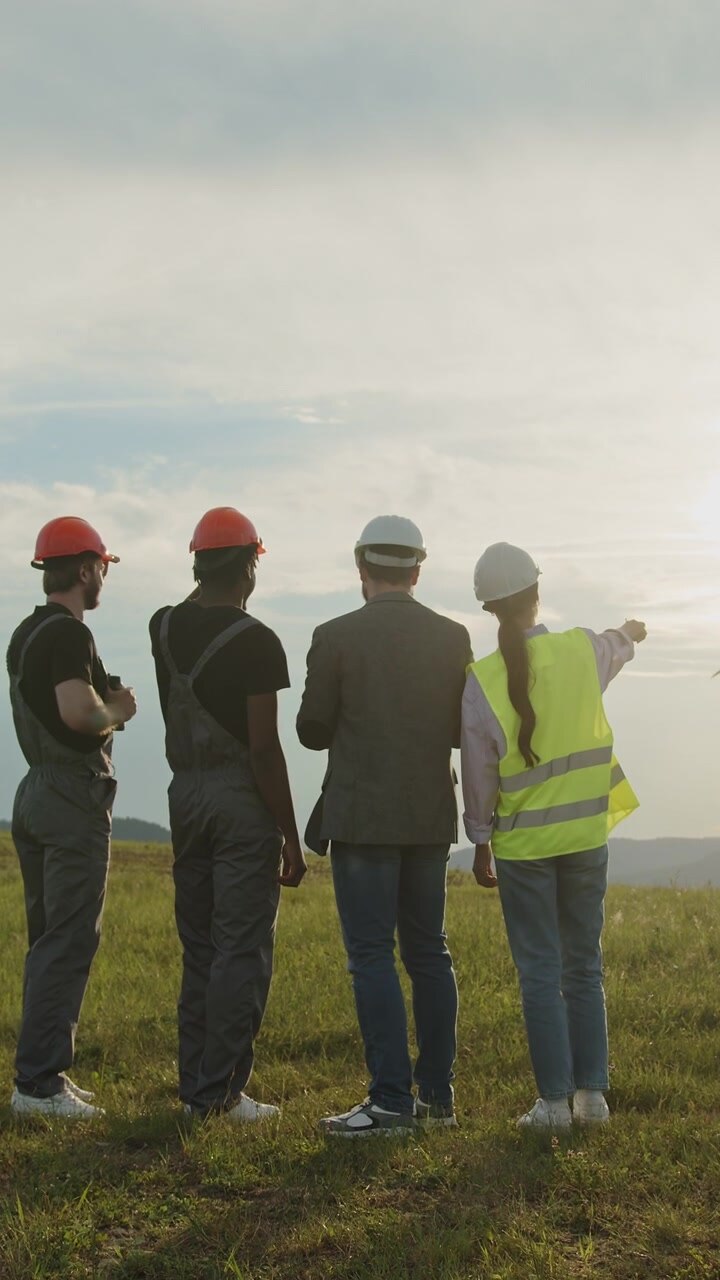 Diverse construction workers in hard hats and vests stand on grassy field discussing plans with