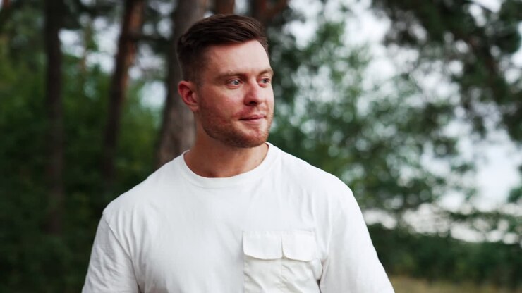 Caucasian unshaved man in white t-shirt walks in the nature. Relaxed man portrait spending time outdoors in summer.