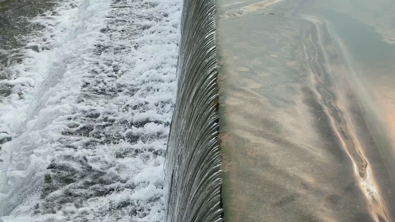 Detailed shot of water spilling over a step dam, contrasting calm surface with foamy turbulence below, highlighting natural flow and engineered structure.