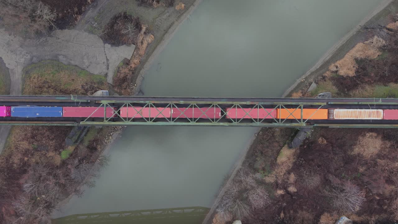 Luftfoto set oppefra af et godstog, der transporterer farverige skibscontainere over en stålbanebro over en rolig flod