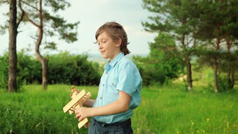 close-up view of happy little boy playing with wooden airplane toy and smiling at camera in the park on a cloudy day