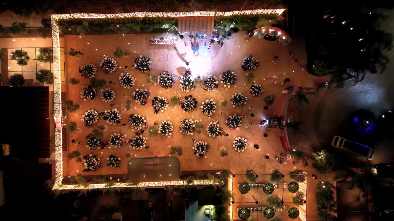 Los invitados se reúnen en un evento gastronómico al aire libre con numerosas mesas dispuestas bajo luces. El ambiente es animado mientras la gente disfruta de sus comidas y socializa por la noche.