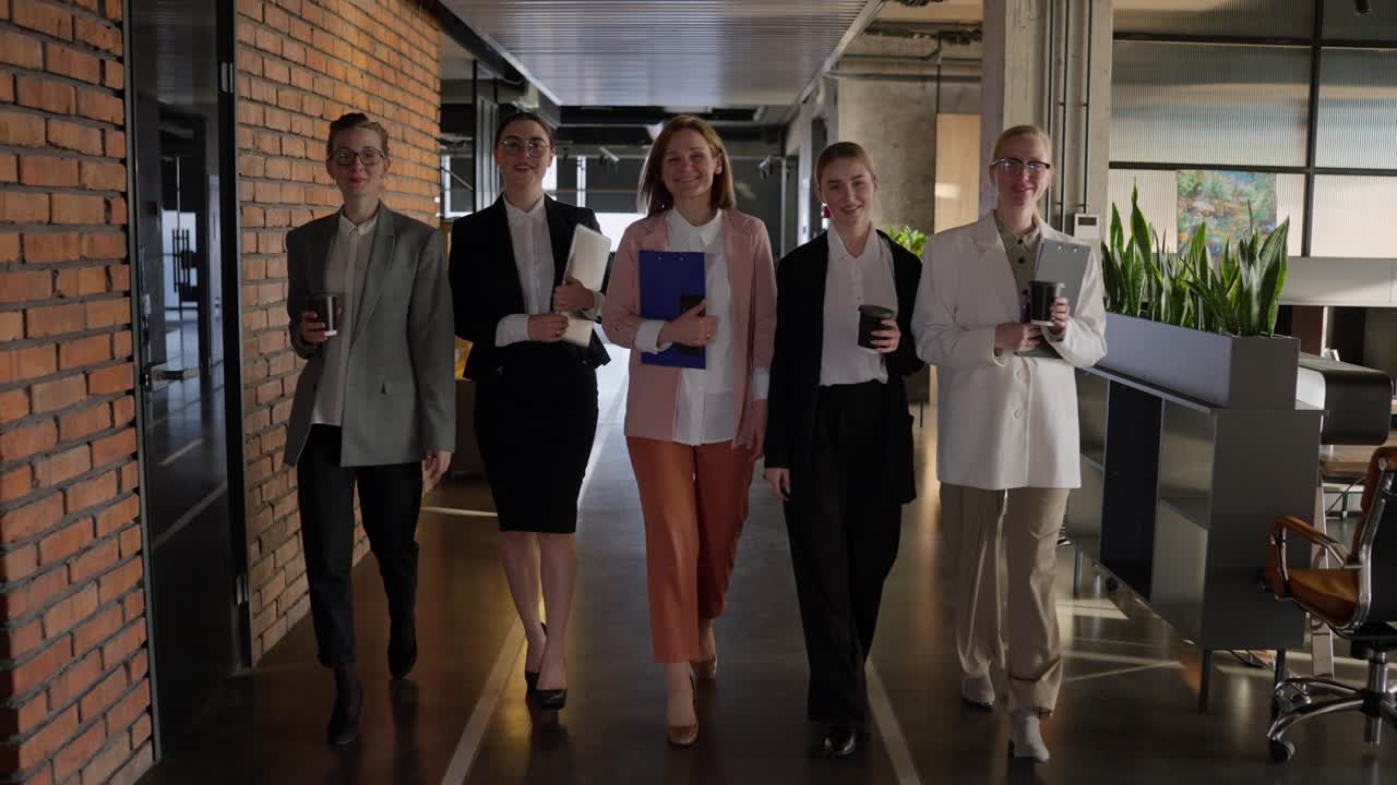 Portrait of a group of five confident businesswomen in business clothes with coffee and laptops in their hands walking along a gray floor in a modern office