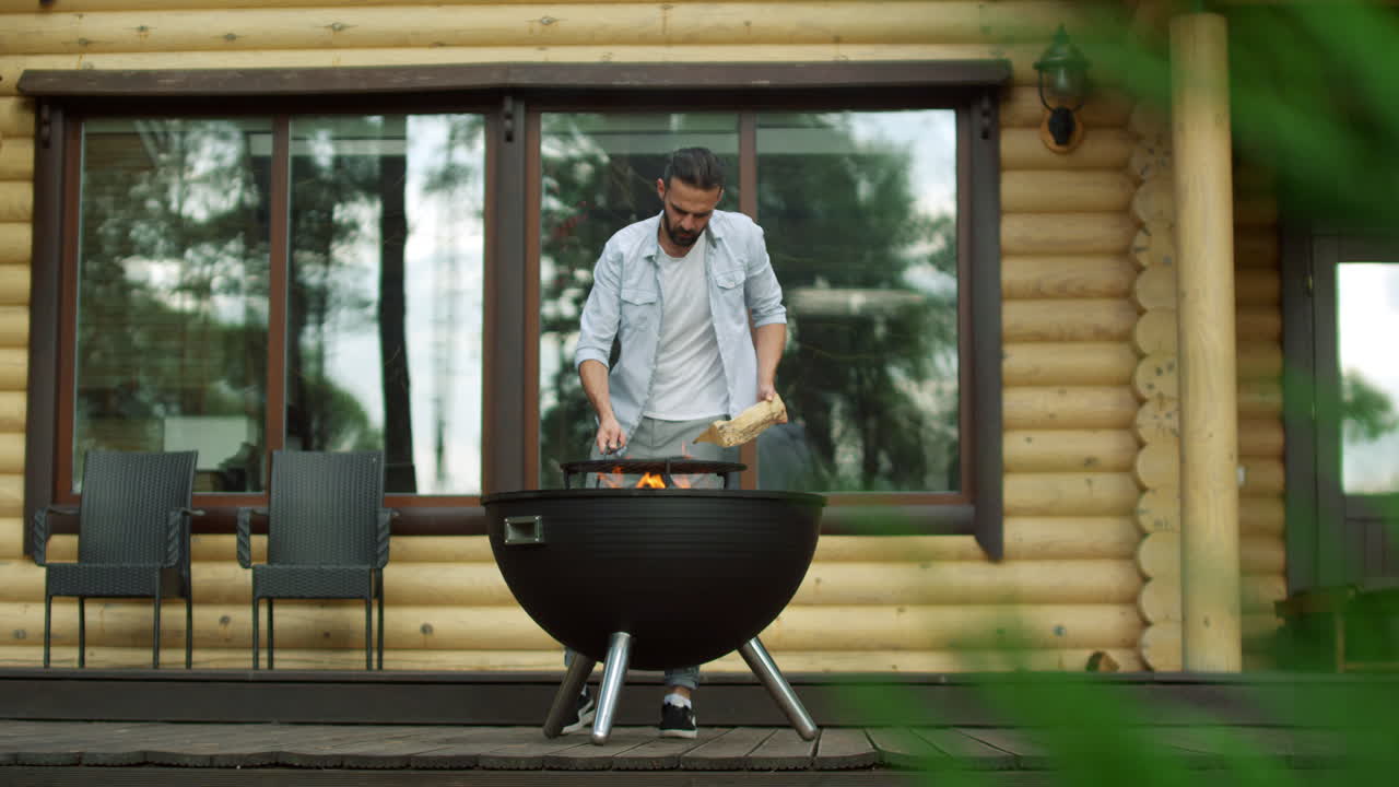 Uomo pensieroso che mette legna da ardere nel barbecue. Ragazzo concentrato che accende il fuoco nella griglia.