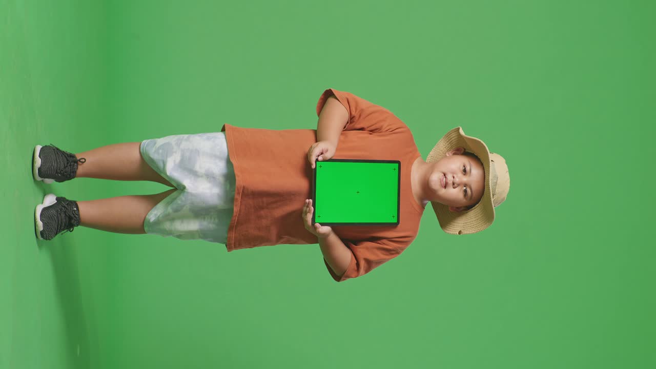 niño turista asiático con un sombrero sonriendo y mostrando la tableta de pantalla verde. niño investigador examina algo en el fondo de la pantalla verde, concepto de aventura de turismo de viaje, cuerpo completo