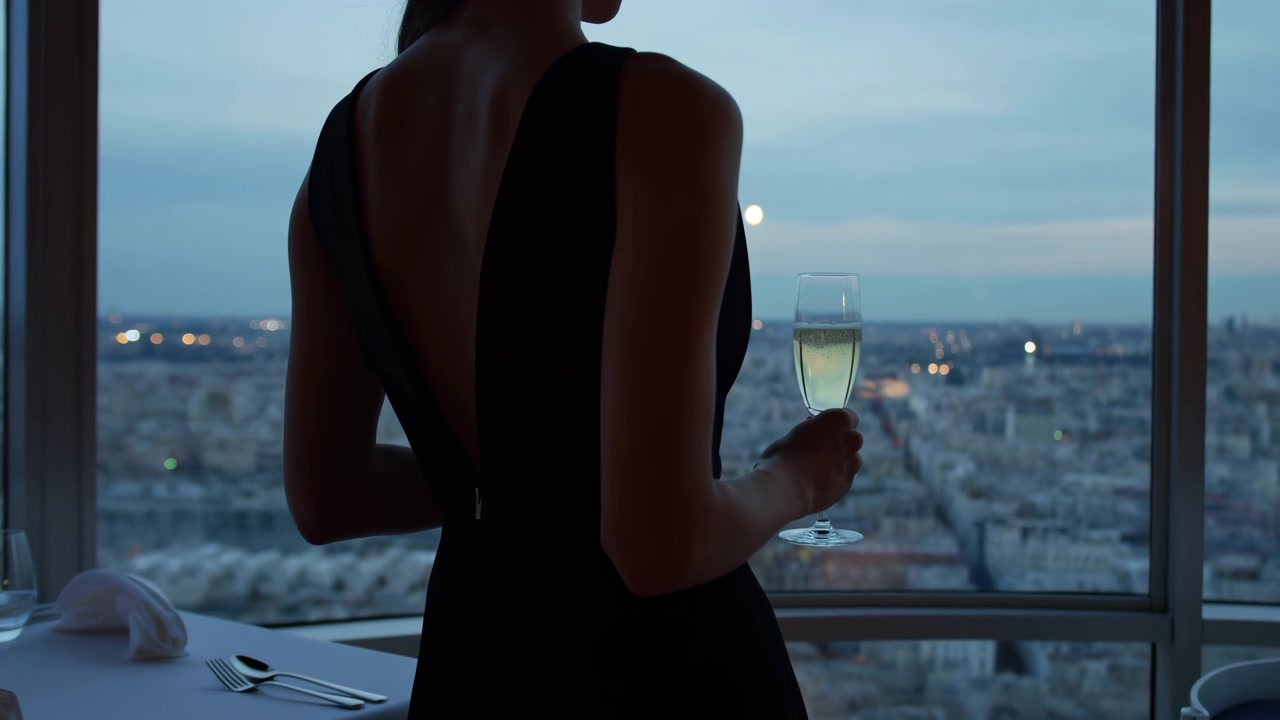 Seen from behind, woman holds a pale beverage near a set dining table while the framing gradually tightens, silhouetting her dark dress against a wide urban view at dusk.