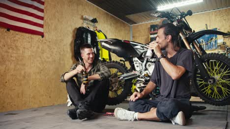 two happy male mechanics drink drinks from small bottles in their garage workshop. break and rest while working in the workshop. auto mechanic duet