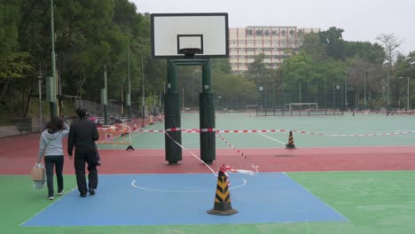 people walk past an empty basketball court seen at a closed playground due to covid-19 coronavirus outbreak and restrictions in hong kong