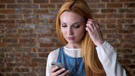 young alluring blond girl is reading message on smartphone, smiling, brick background, communication concept