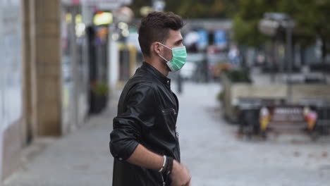 un joven genial con una chaqueta de cuero sintético con un peinado moderno en una calle concurrida y se pone una máscara protectora verde covid-19, luego se mete las manos en los bolsillos, de lado a la cámara, 4k
