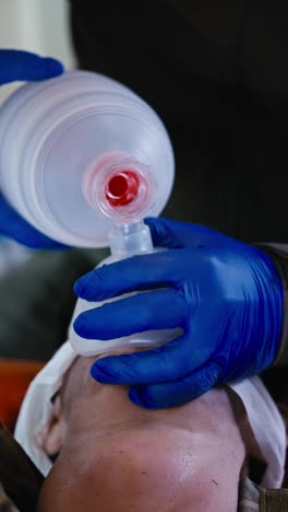 Vertical-video-close-up-of-a-confident-man-in-blue-medical-gloves-pumping-air-and-performing-artificial-respiration-on-a-man-who-is-lying-on-a-couch-in-a-small-medical-room