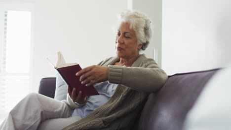 Eine-Afroamerikanische-Seniorin-Liest-Ein-Buch,-Während-Sie-Zu-Hause-Auf-Der-Couch-Sitzt