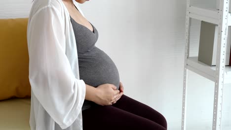 mujer embarazada tocando su estómago con la mano suavemente mientras está sentada en el sofá cerca de la ventana en el dormitorio de la casa. estilos de vida y atención médica de la gente. bebé recién nacido y maternidad. concepto de madre y madre soltera.