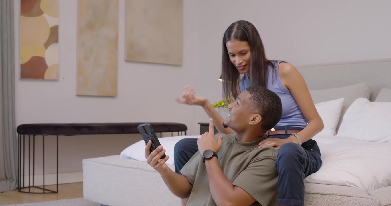 Couple man sitting on floor holding phone showing content woman leaning and pointing on bed smiling