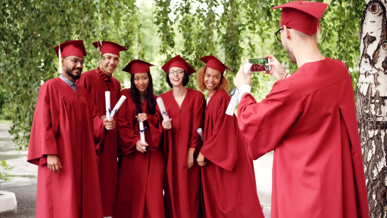 Cheerful graduating students are standing in line outdoors and young man with smartphone is shooting them while graduates are laughing, posing, waving hands.