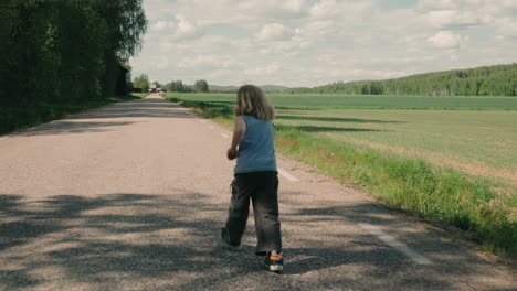 young child runs along asphalt country road next to farm fields, rear tracking