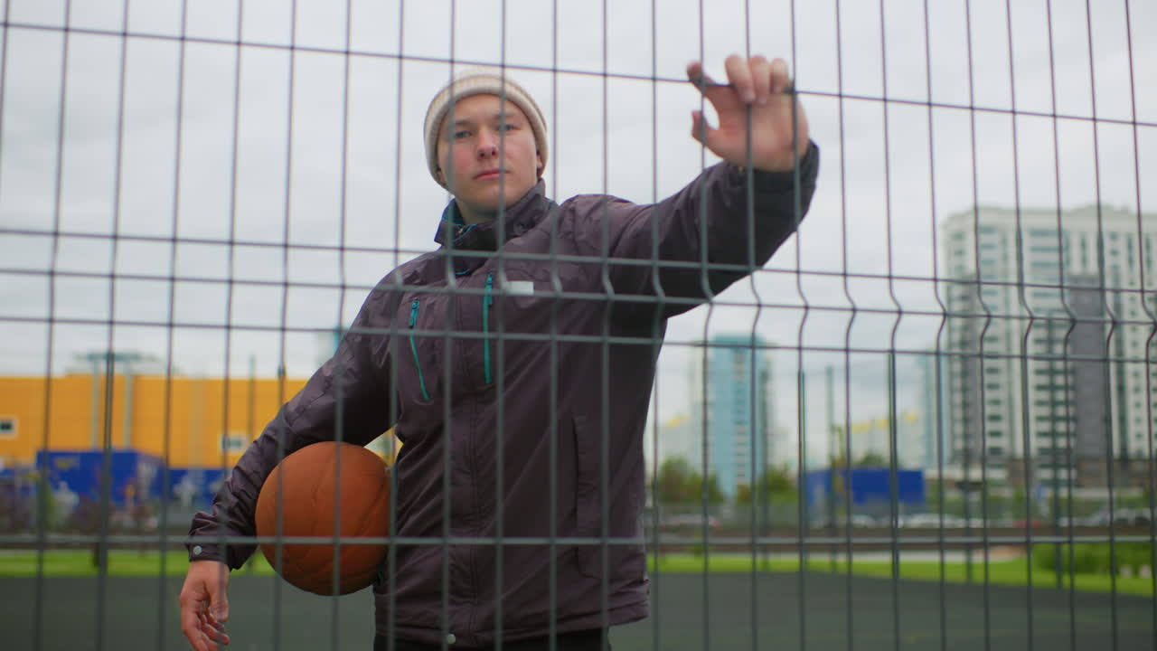Man with basketball behind wire fence, standing on outdoor urban court under overcast sky, wearing hoodie and beanie, holding ball at hip, reaching through mesh toward camera, high rise buildings