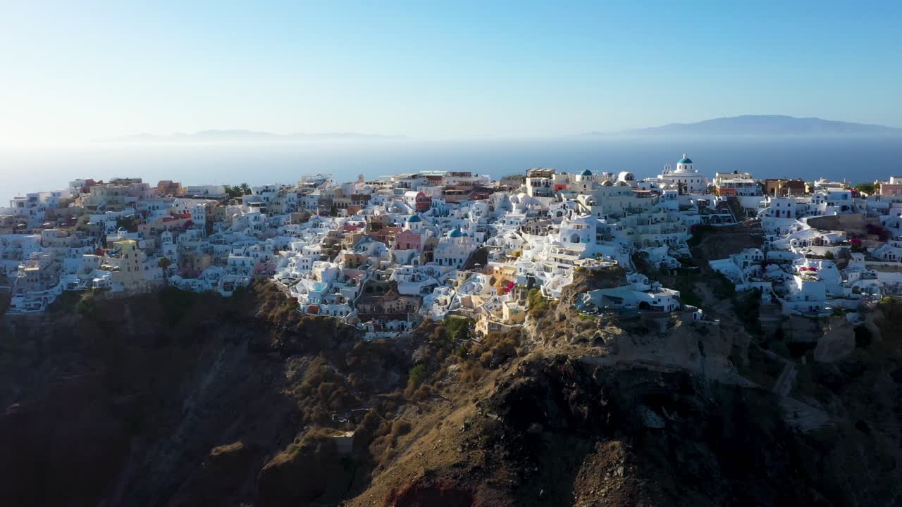 Drone flying sideways alongside famous village Oia with white cave houses, villas and caldera in Santorini, Greece.