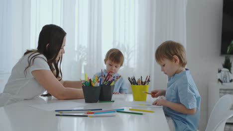 mom helps two sons to perform preschool homework to draw a pencil drawing sitting at the table in the house