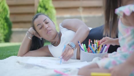 una madre y su hija pequeña se sientan en una manta en el patio trasero, participando en una actividad de dibujo juntos. la escena al aire libre captura un momento familiar pacífico, fomentando la creatividad y el vínculo.