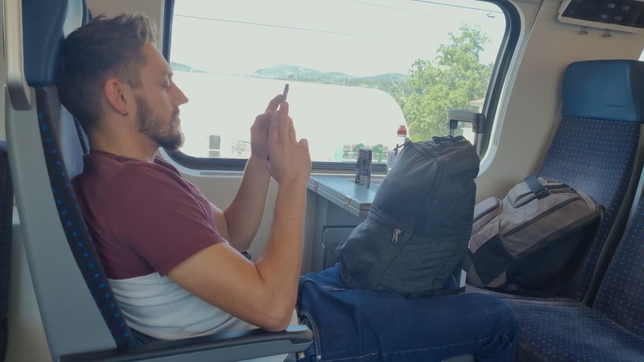 A man sits relaxed on a Swiss train with his luggage, scrolling on his phone as Swiss buildings and a spring landscape pass by outside the window