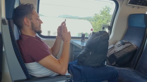 a man sits relaxed on a swiss train with his luggage, scrolling on his phone as swiss buildings and a spring landscape pass by outside the window