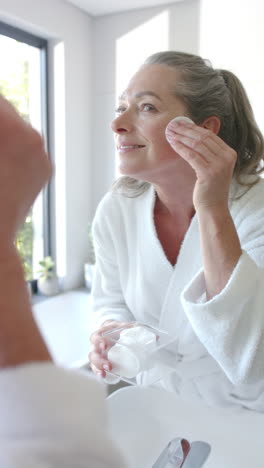 vertical video: applying skincare, woman in bathrobe using cotton pad in bathroom mirror
