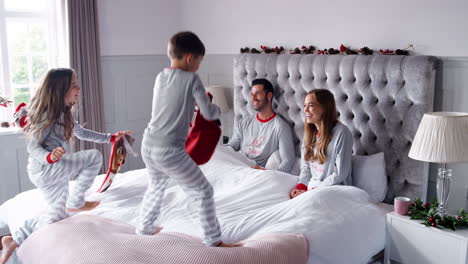Excited-Children-Running-Into-Parents-Bedroom-At-Home-With-Stockings-And-Bouncing-On-Bed-As-Family-Open-Gifts-On-Christmas-Day