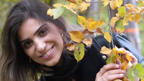 brunette girl in autumn forest is holding a branch with yellow leaves and hiding behind it, smiling at camera