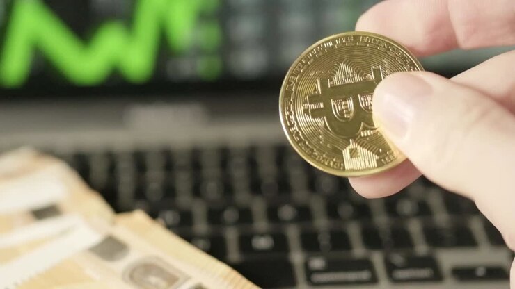 Primo piano di dita che giocano con una moneta d'oro Bitcoin, grafico di criptovaluta verde in crescita, con laptop sullo sfondo e schermo che mostra banconote in euro