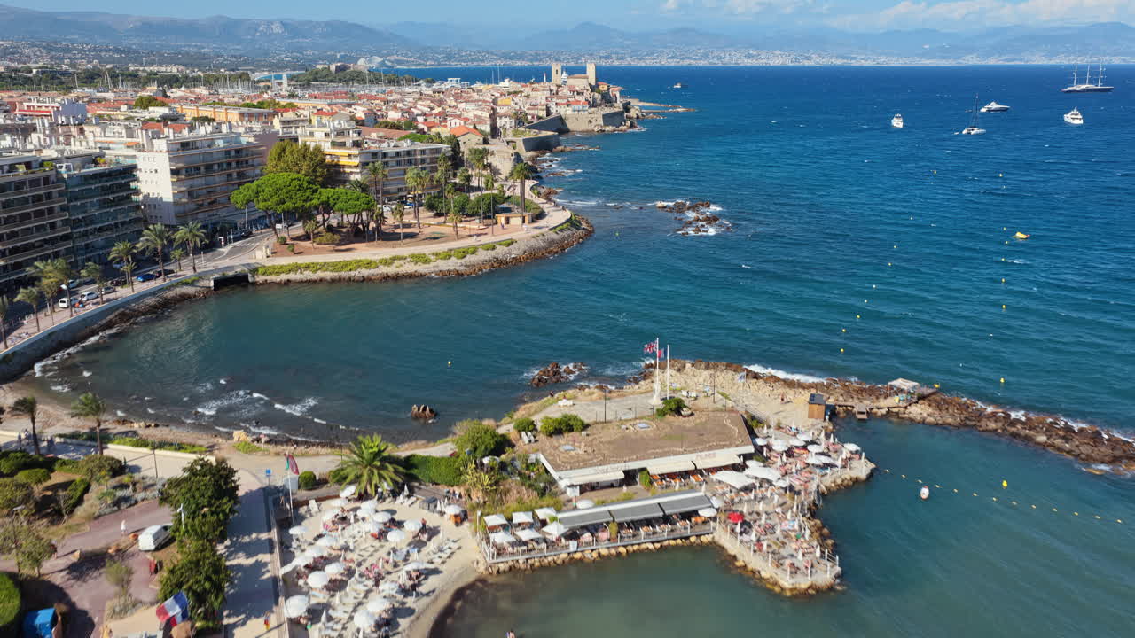 Aerial drone view of Nice's sandy beach filled with sunbathers and swimmers, with palm lined Promenade des Anglais and seafront buildings behind