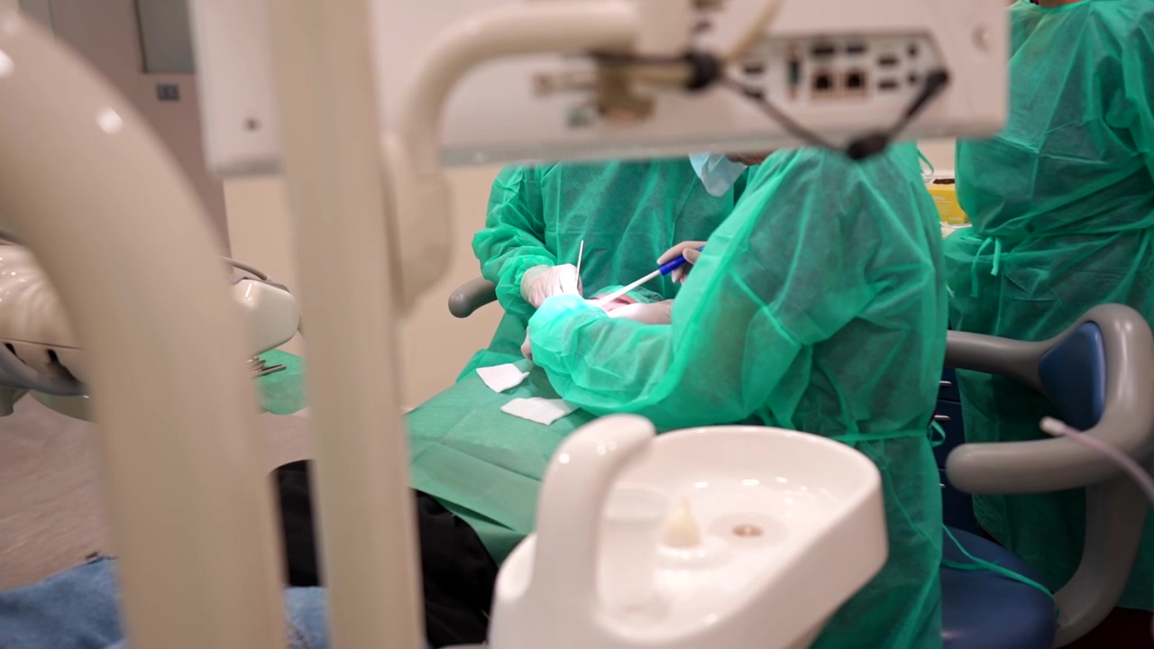 Odontólogos irreconocibles trabajando con un paciente durante una cirugía de injerto dental, vistos desde detrás del sillón dental.