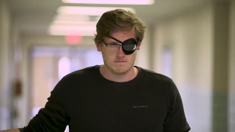 white man with eye patch and glasses stares at camera seriously in a hallway