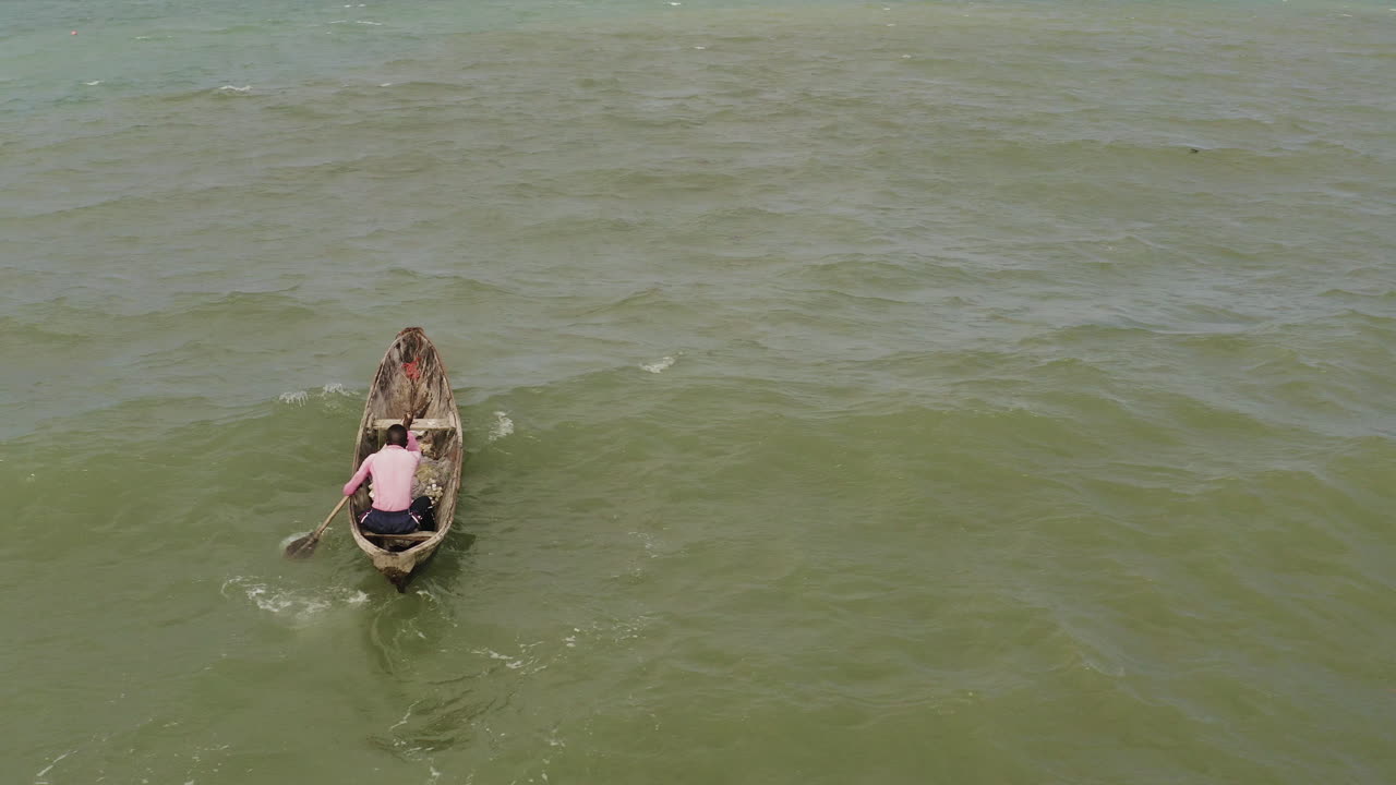 A man paddling a wooden canoe boat against waves and wind near the coast of Dar es Salaam on a sunny day, Tanzania