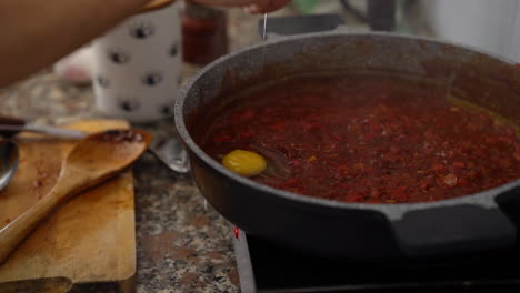 man-breaks-an-egg-into-a-bubbling-tomato-sauce,-preparation-of-homemade-Shakshouka,-filmed-in-slow-motion-and-natural-day-light