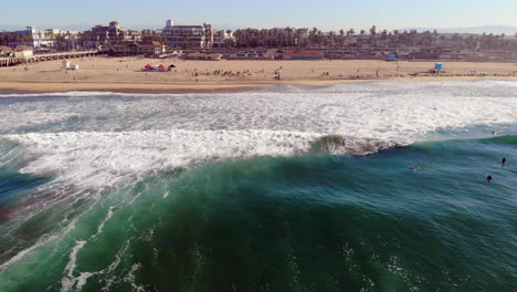 wide aerial 4k drone boom down of huntington beach shore from over ocean waves, looking back at downtown and beach sand with surf in foreground at sunrise, pacific ocean, southern california