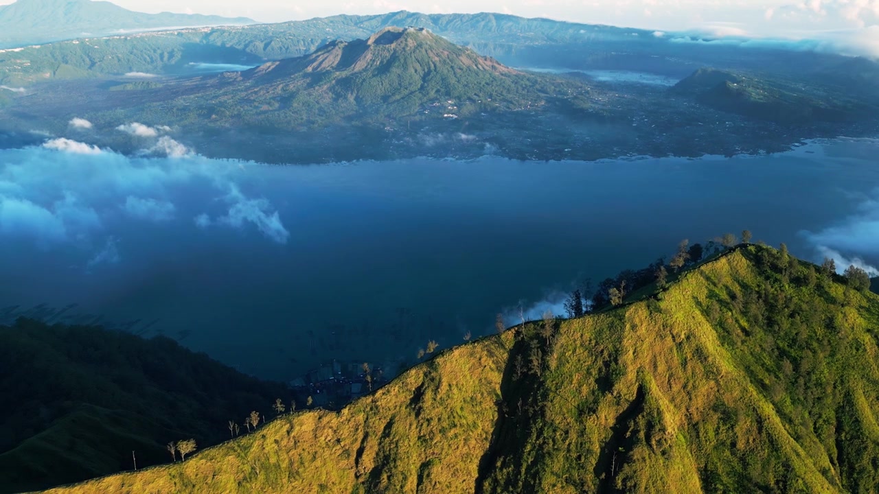 Lago di caldera vulcanica in Indonesia con pendii montuosi illuminati dal sole e nuvole, il Monte Batur visibile sullo sfondo, riprese aeree con drone.