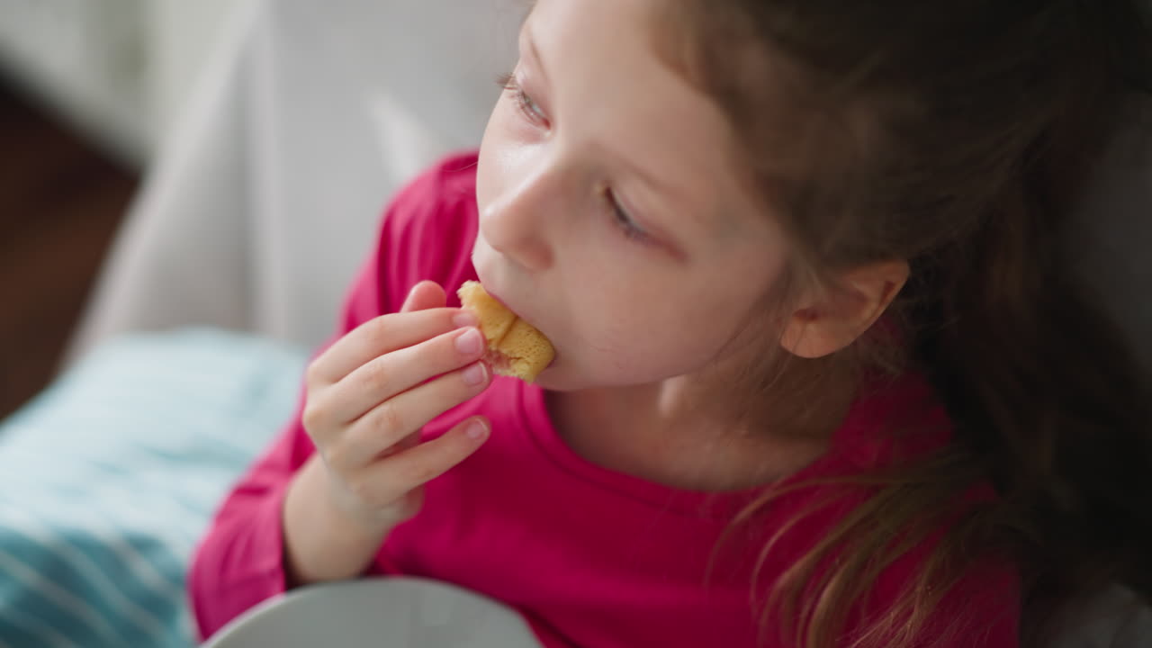 Overhead view of little girl in pink top forcing entire dessert into mouth with determined expression while sitting on bed with soft light background
