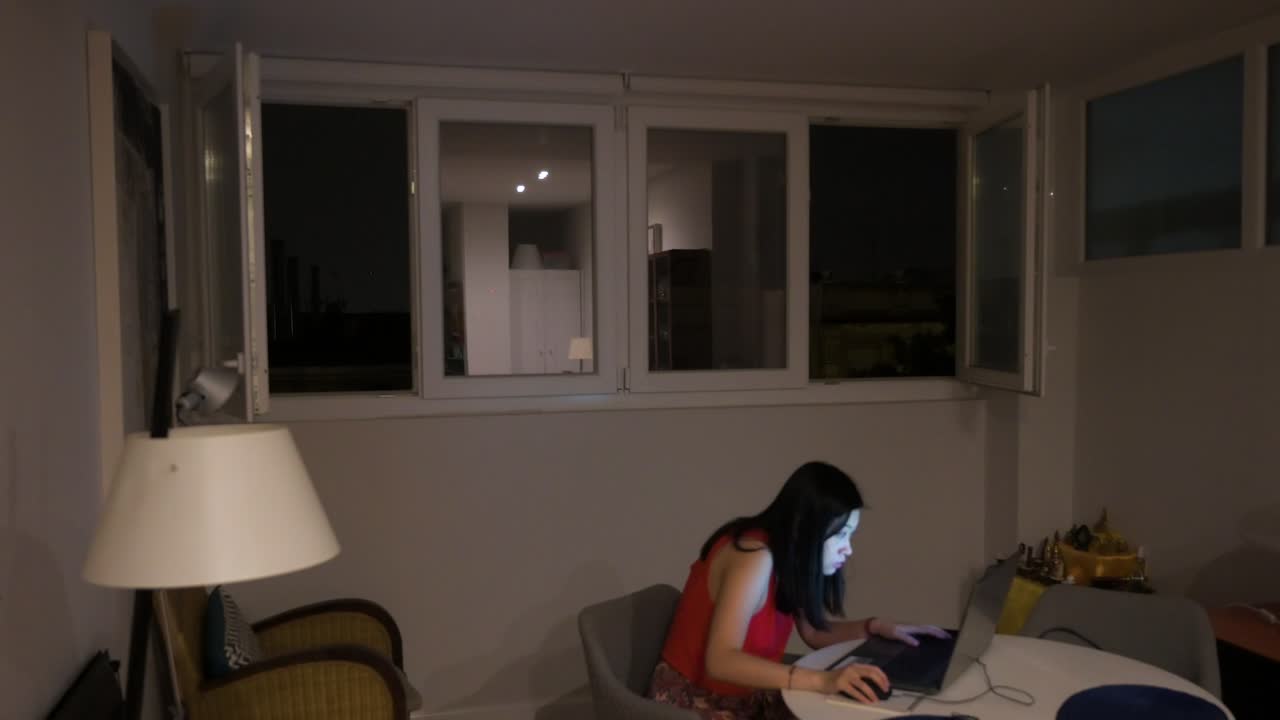 Una donna usa un laptop al chiuso mentre fuori c'è un temporale, con lampi visibili attraverso le finestre di casa