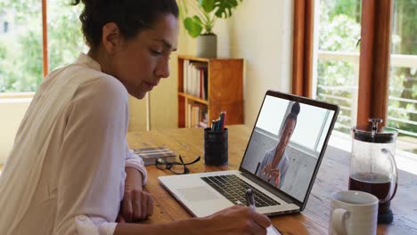 mujer afroamericana tomando notas mientras tiene una videollamada en una computadora portátil en casa