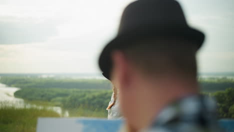 a woman in a white dress and hat poses gracefully in a grassy field overlooking a serene lake, with a blurred close-up of a man in a hat in the foreground