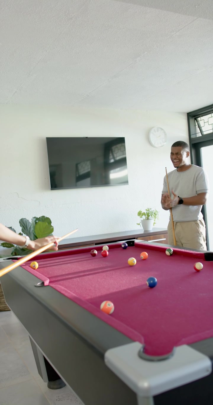 Vertical video: Diverse duo playing pool for fun at home, left player striking cue, friend cheering