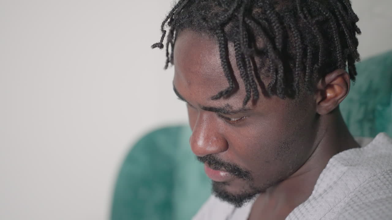 Close-up of man in white bathrobe deeply focused on reading black notebook, looking down with concentration, soft lighting, green velvet headboard in background, serene indoor setting
