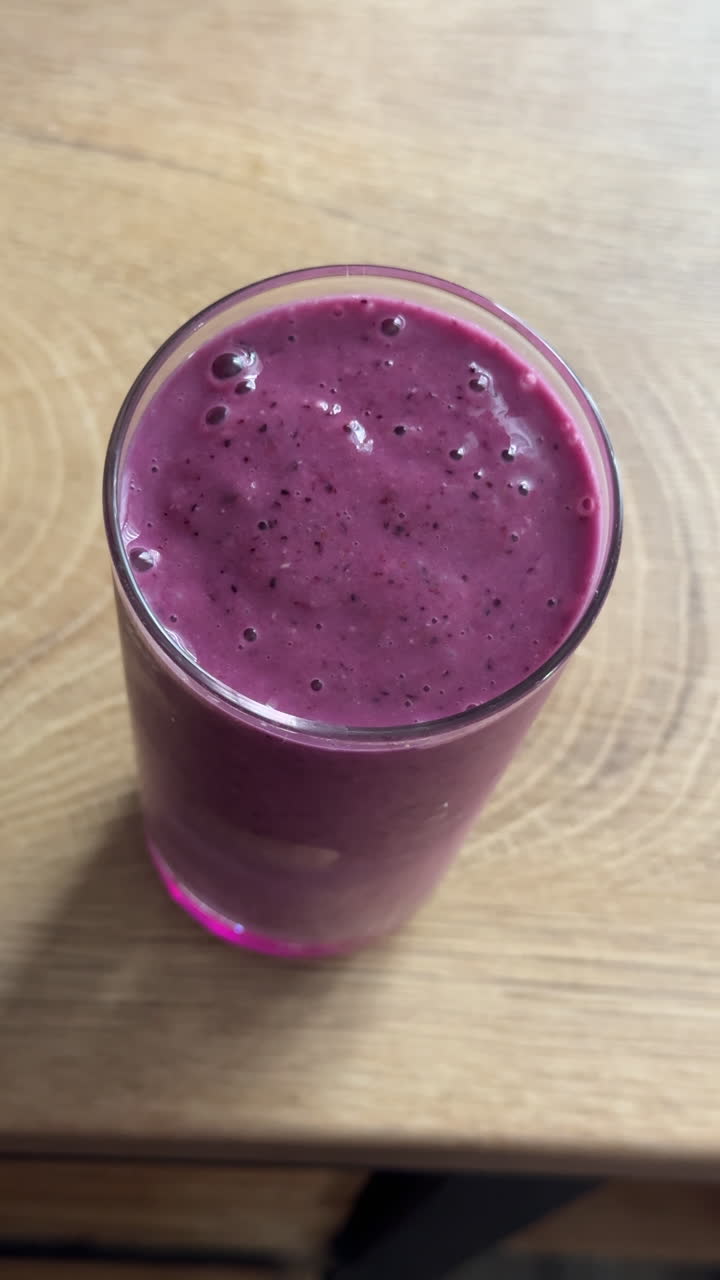 Scatto verticale in stile UGC di una mano che prende un bicchiere pieno di denso frullato di frutti di bosco su un tavolo di legno. Bevanda di frutta naturale fatta in casa con consistenza cremosa e colore vivace.